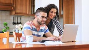A guy and girl looking at a laptop in their kitchen. 